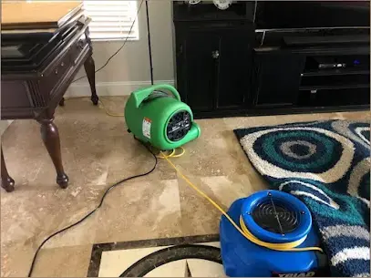 Air movers drying a stone floor and rug beside a piano after a leak
