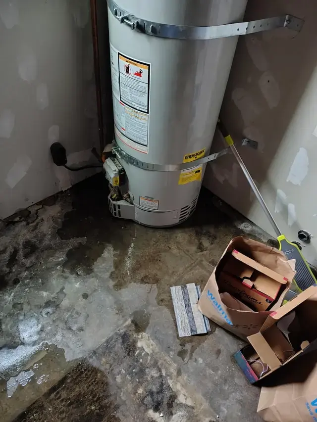 Water heater in a garage corner with water and debris across the floor